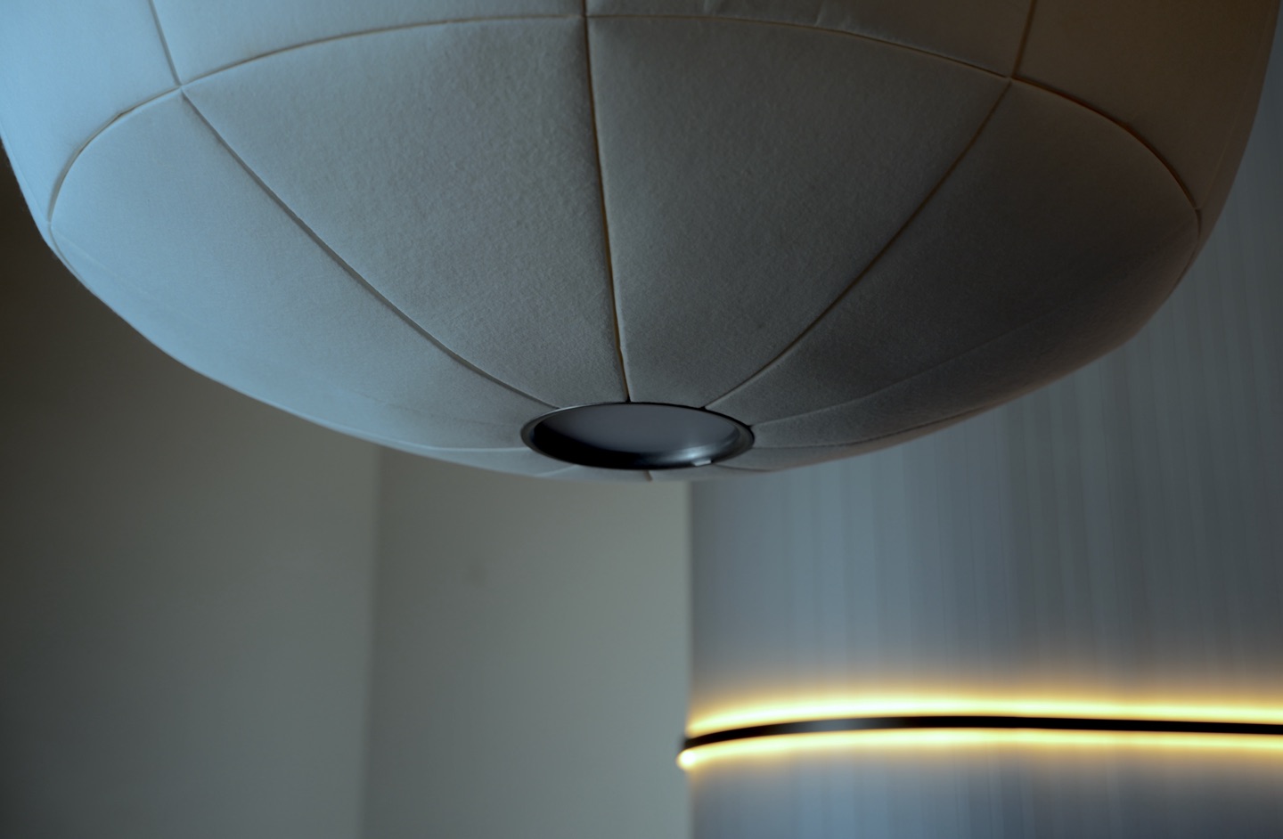 Underside of an unlit wool pendant, radial seams meeting at a metal trim ring, with a lit band on a white column behind.