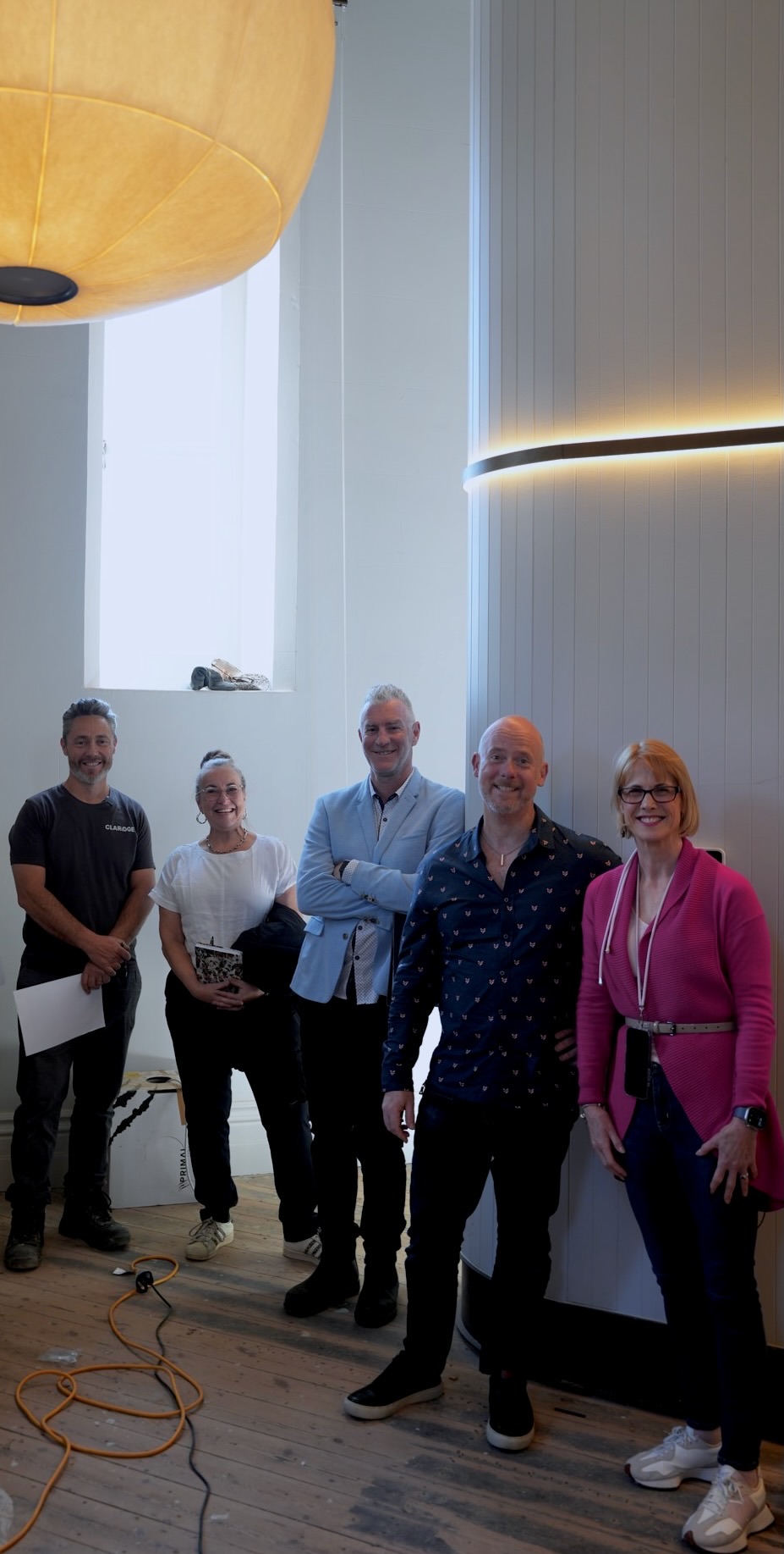 Mark Di Mauro, Bobbie-Jane Skewes, Mike Sheidow, Daniel Johnson and Michelle Simper stand together in a tower bedroom beneath a lit wool pendant, a tall narrow window behind them and a light strip curving along the wall.