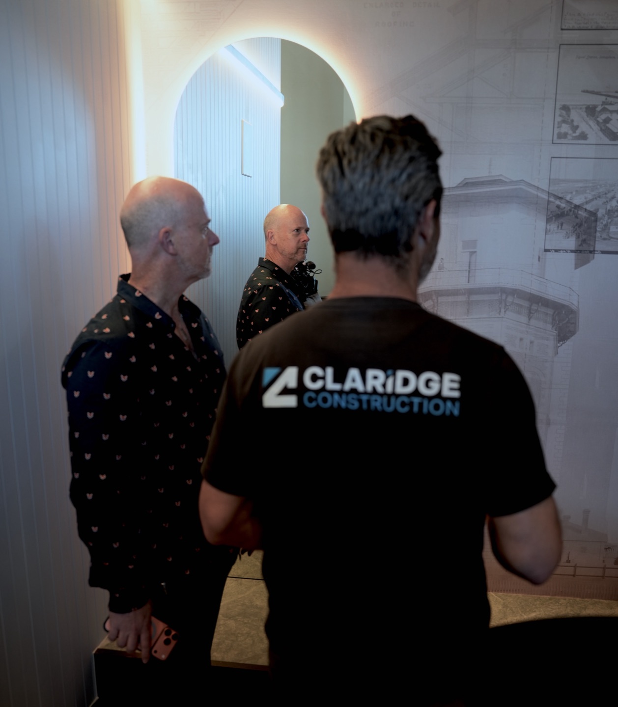 Mark Di Mauro, in a Claridge Construction shirt, and Daniel Johnson at a lit arched mirror, Daniel reflected in it, with the tower's heritage drawings on the wall behind.