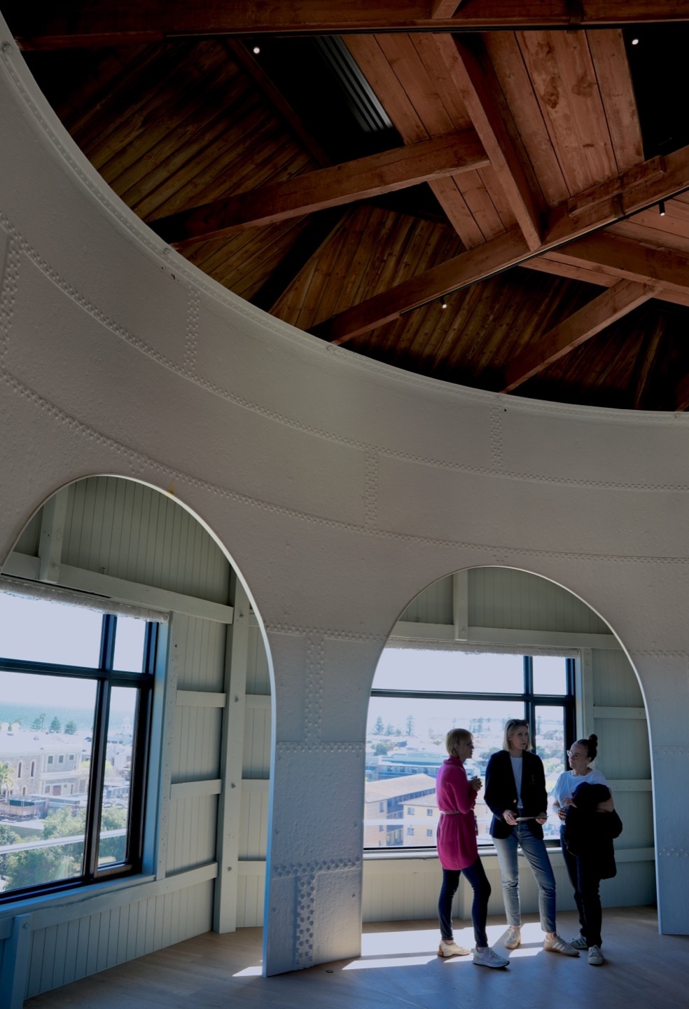 Inside the Observatory at the top of the tower: Michelle Simper, Emma Blomfield and Bobbie-Jane Skewes stand in an arched window opening beneath the timber roof structure, with Semaphore's rooftops and the gulf behind them.