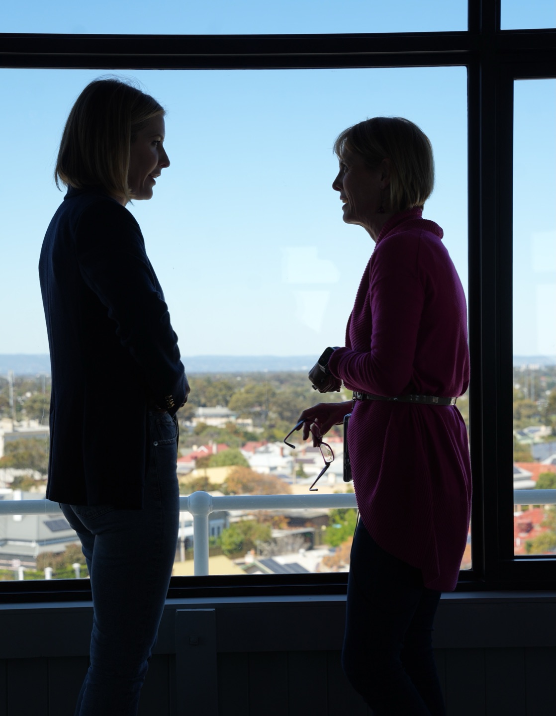 Emma Blomfield and Michelle Simper in silhouette, talking at the Observatory's window, the rooftops of Semaphore stretching to the hills behind them.