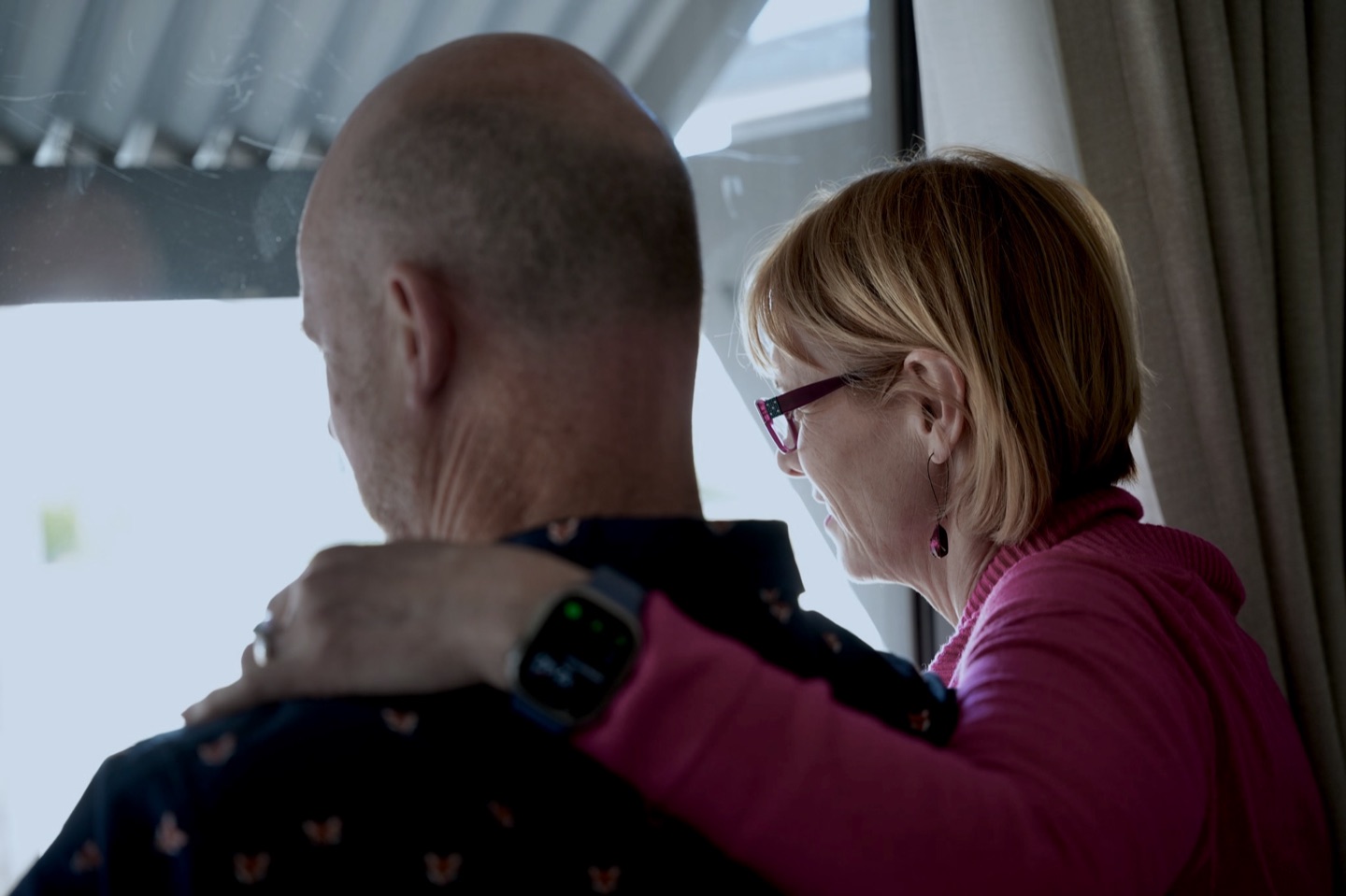 Seen from behind, Daniel Johnson and Michelle Simper look out through a Retreat window past its curtain, her arm around his shoulder.